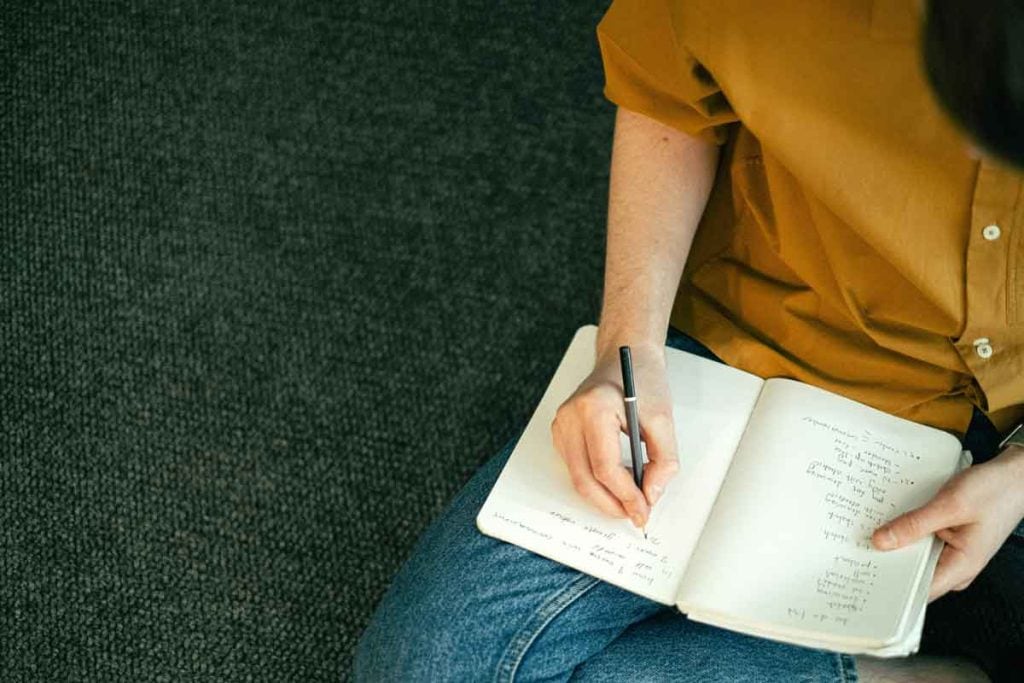 Marketing clichés can help you create memorable brand messaging, like this person wearing a gold shirt and jeans, writing in a notebook with a pencil.